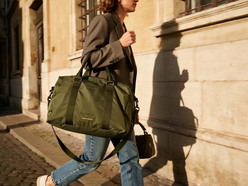 Woman walking on a cobblestone European street carrying an olive travel tote and crossbody.